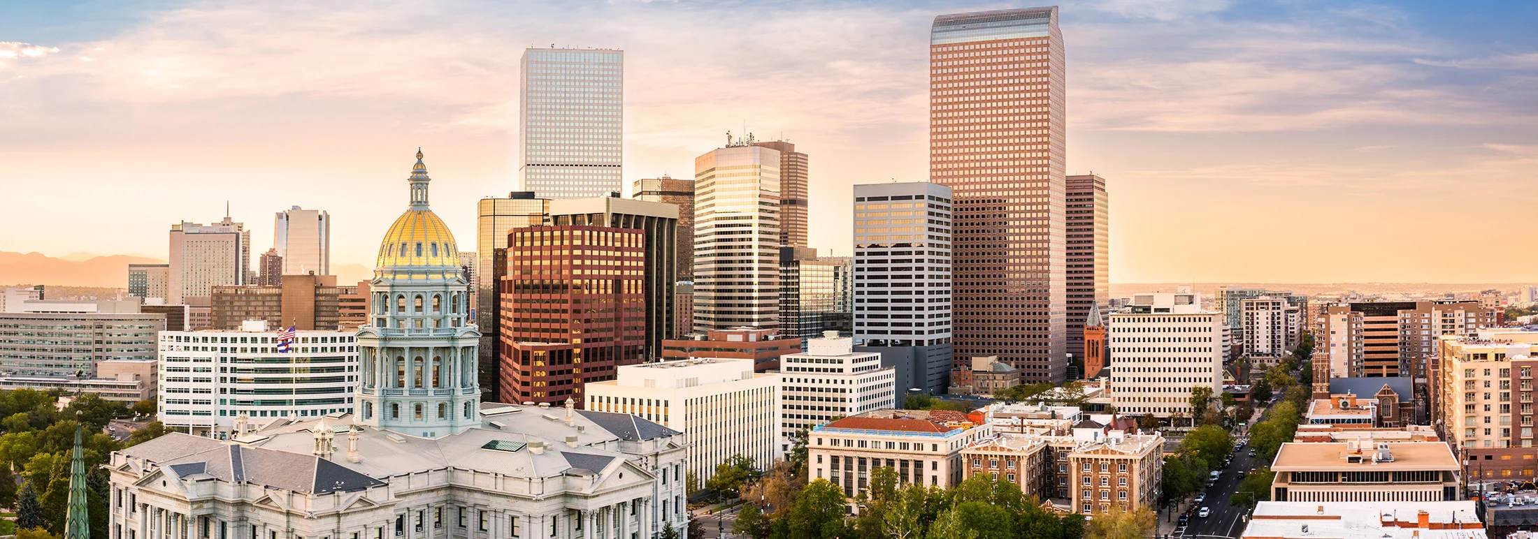 Downtown Denver skyline with the Colorado State Capitol’s gold dome in the foreground, surrounded by modern office towers under a soft, pastel sunset sky.