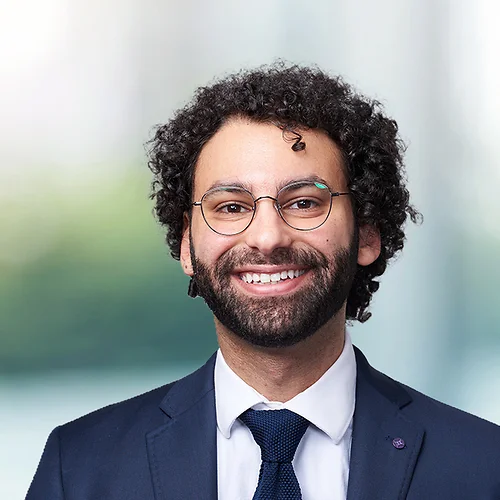 A smiling man with curly dark hair and a beard, wearing glasses, a navy suit, white shirt, and tie, posed against a softly blurred office background.
