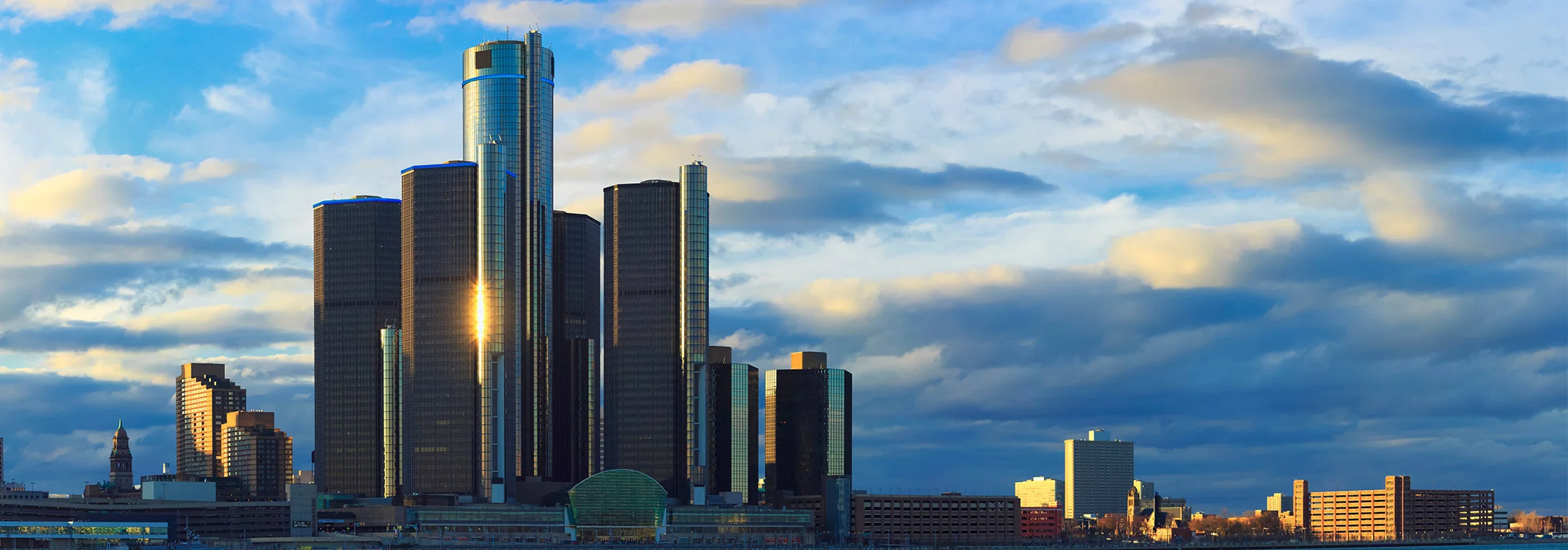 Detroit skyline at sunset featuring the Renaissance Center’s cylindrical glass towers along the Detroit River, with dramatic clouds and warm light peaking through the buildings.