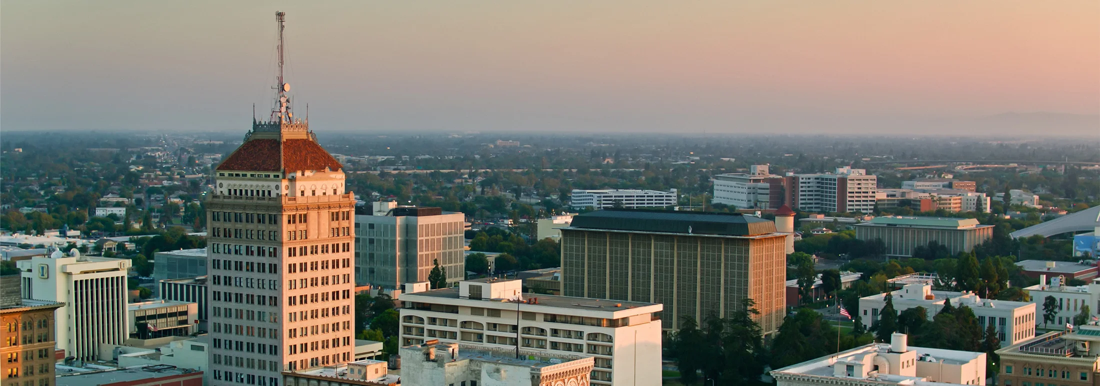 Fresno city skyline at dusk highlighting a high-rise building with a red-tiled roof and rooftop antennas, surrounded by low-rise offices and trees stretching into the distance.