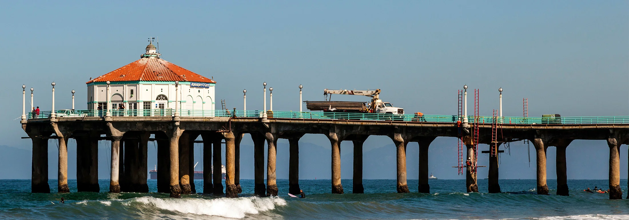 Hermosa Beach pier extending over the Pacific Ocean with a round pavilion topped by a red tile roof, ocean waves breaking below, and a clear blue sky in the background.