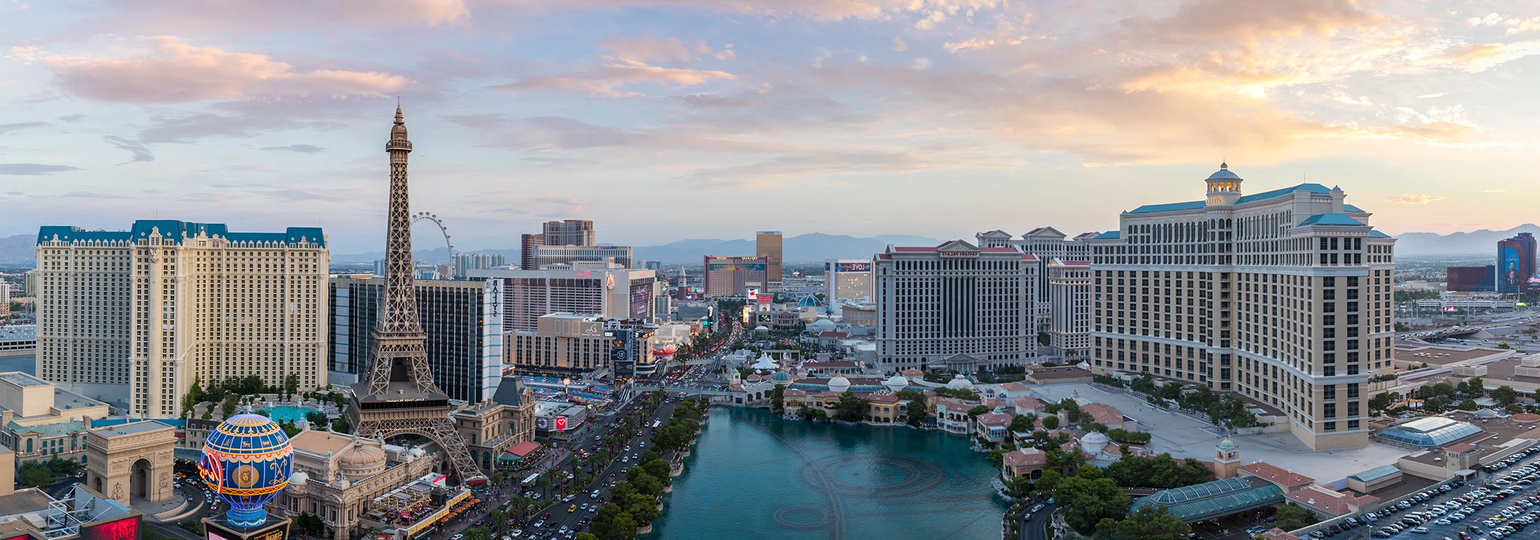 Panoramic view of the Las Vegas Strip at sunset, featuring the Paris Las Vegas Eiffel Tower replica, the Bellagio fountains and lake, and surrounding resort hotels under a soft, glowing sky.