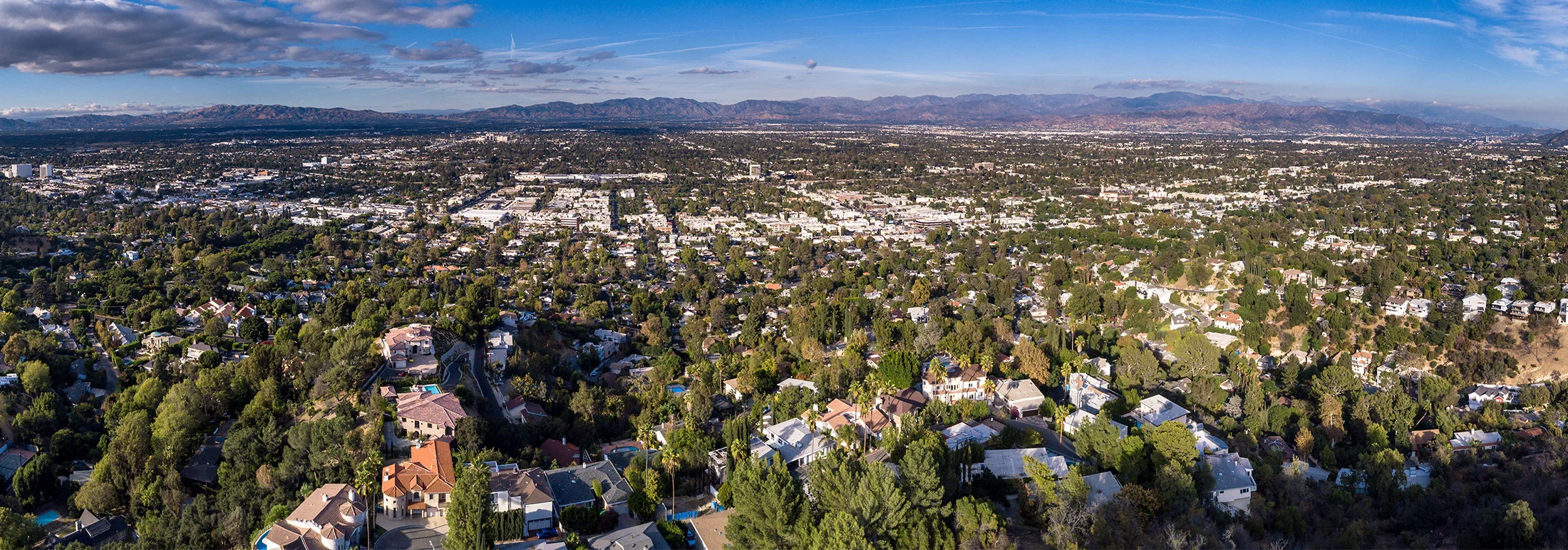 Aerial panoramic view of Woodland Hills, California, showing a sprawling residential neighborhood with tree-covered streets and homes stretching toward distant mountains beneath a clear blue sky.
