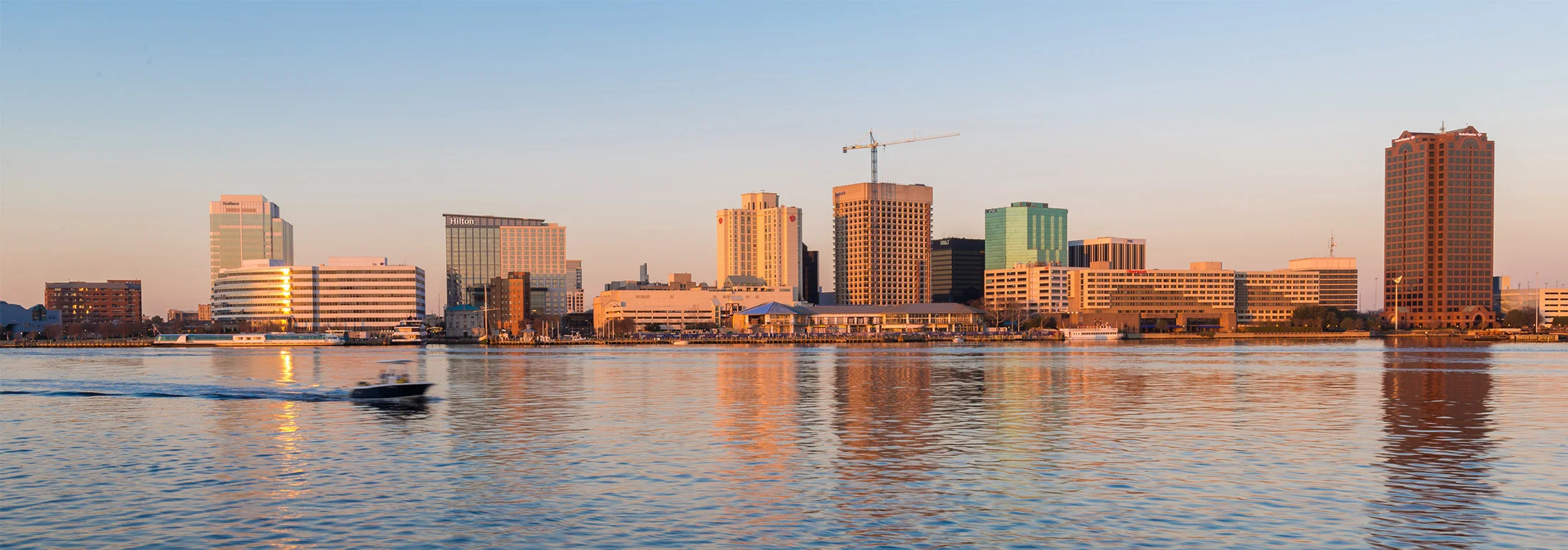 Waterfront skyline of downtown Norfolk at golden hour, showing modern office buildings and hotels reflected in calm harbor water with a small boat passing in the foreground.