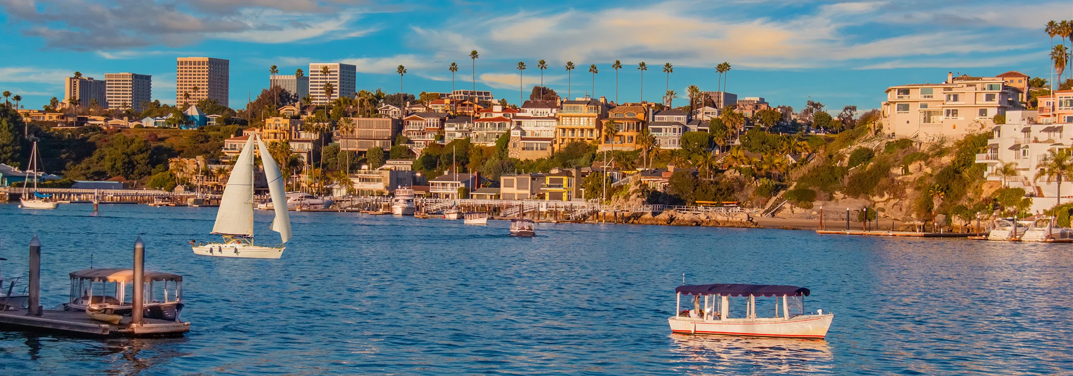 Coastal harbor scene in Orange County, California, featuring sailboats and small boats on calm blue water, hillside homes and palm trees along the shoreline, and warm sunset light illuminating the neighborhood.