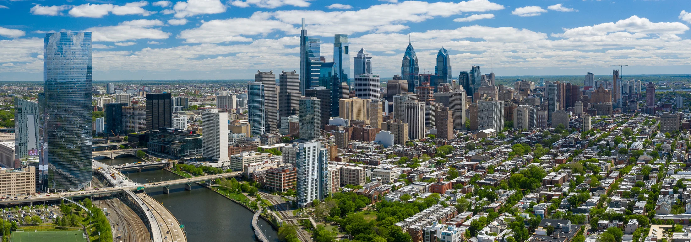 Panoramic aerial view of downtown Philadelphia, showing dense city blocks, modern skyscrapers including glass towers, and the Schuylkill River with bridges curving through the foreground under a partly cloudy blue sky.