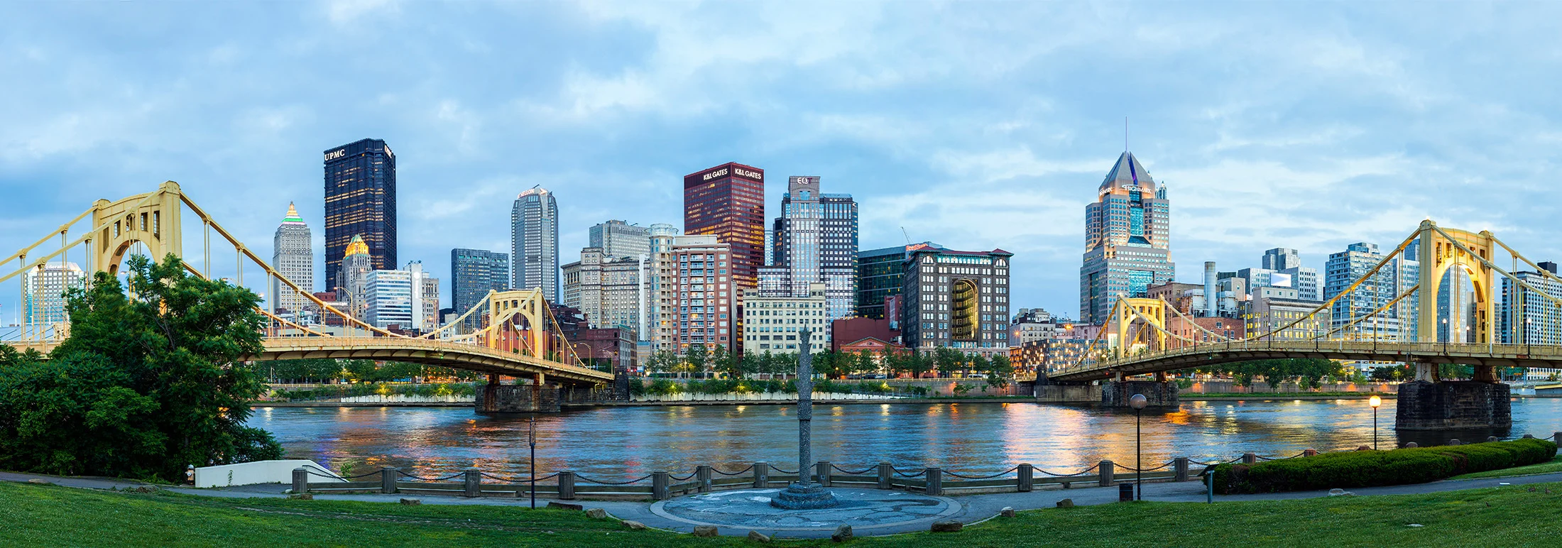 Wide view of downtown Pittsburgh at dusk, with yellow suspension bridges crossing a river in the foreground and a lit skyline of high-rise buildings reflected in the water beneath an overcast evening sky.