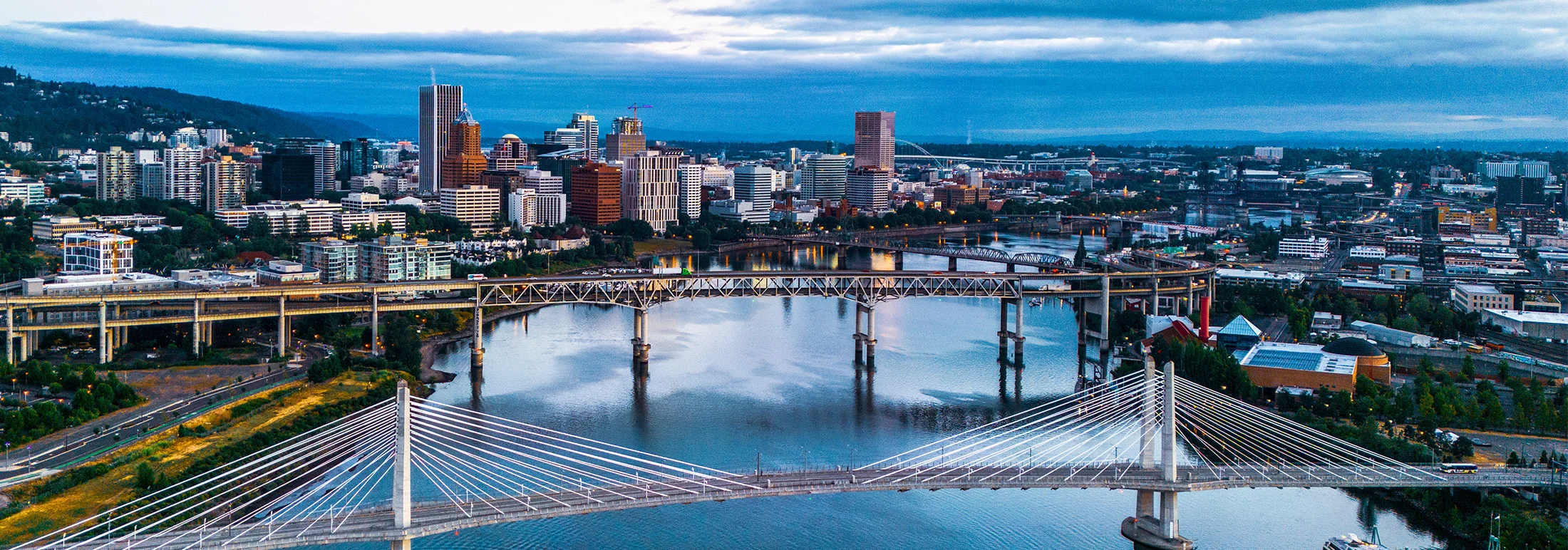 Panoramic view of downtown Portland, Oregon, featuring bridges spanning the Willamette River in the foreground and a dense skyline of modern and historic high-rise buildings under an overcast blue-gray sky.