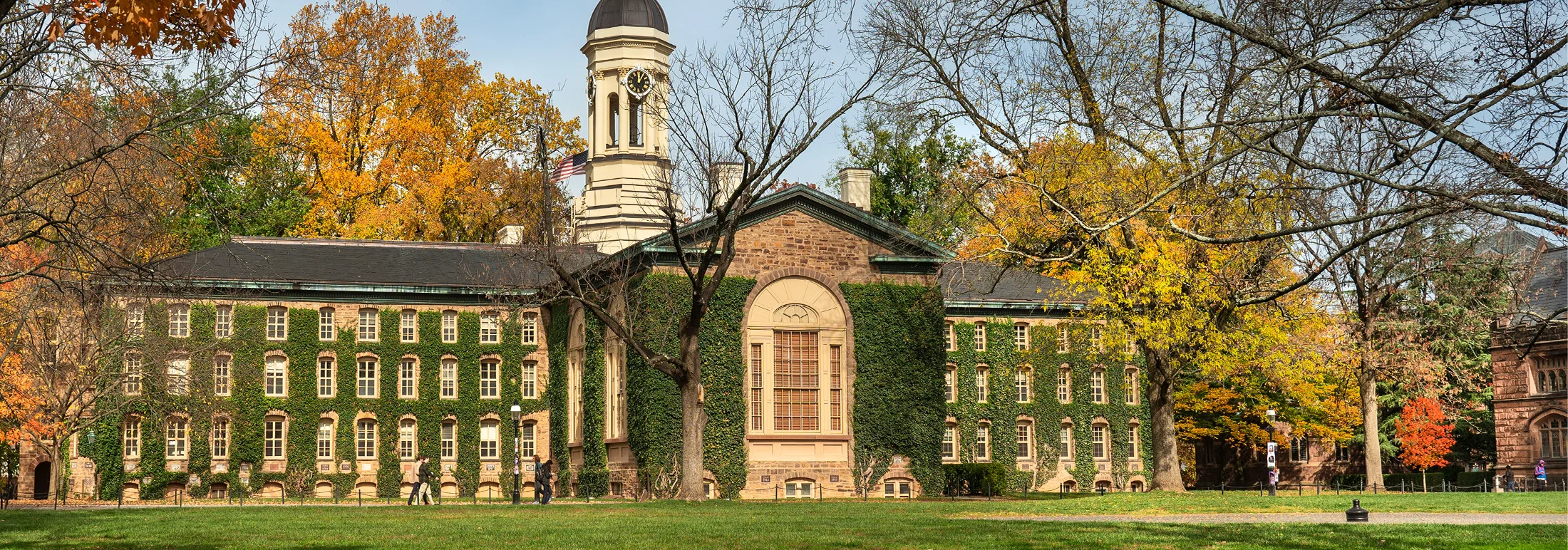 Historic stone academic building at Princeton University covered in ivy, with a clock tower and arched windows, surrounded by autumn trees with yellow and orange leaves and a grassy lawn in the foreground.