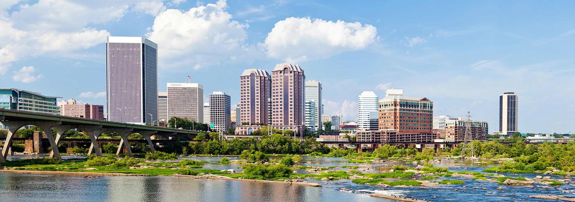 Wide view of downtown Richmond, Virginia, with mid- and high-rise office buildings rising behind a river filled with rocks and greenery, and a concrete bridge crossing from the left under a bright, partly cloudy sky.