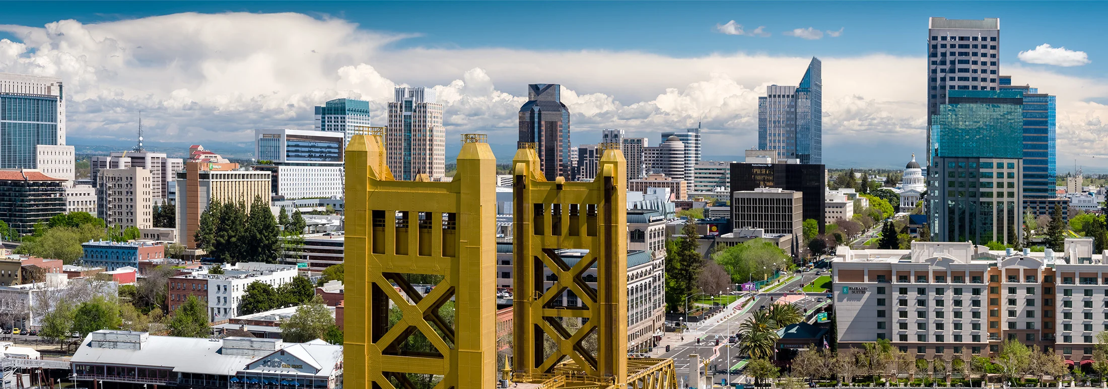 Downtown Sacramento skyline with the golden towers of the Tower Bridge in the foreground, surrounded by modern office buildings and tree-lined streets under a bright sky with large white clouds.