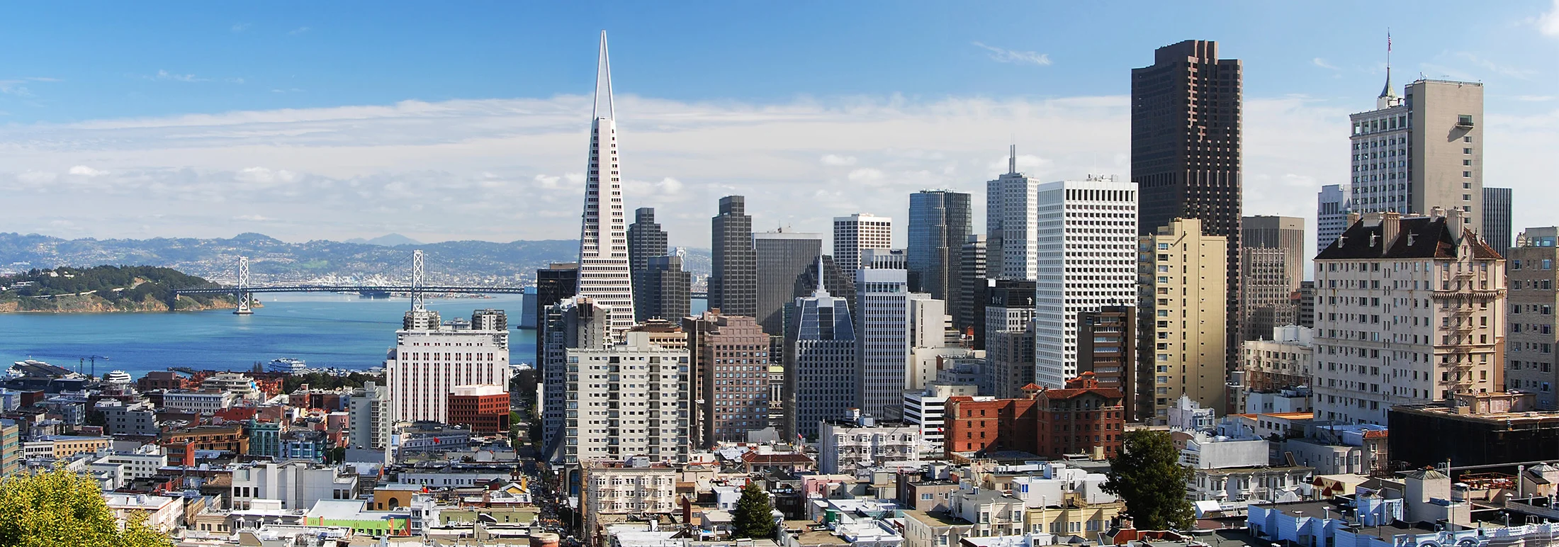 Wide view of downtown San Francisco showing the Transamerica Pyramid among dense high-rise buildings, with the San Francisco–Oakland Bay Bridge and blue bay water visible in the background.