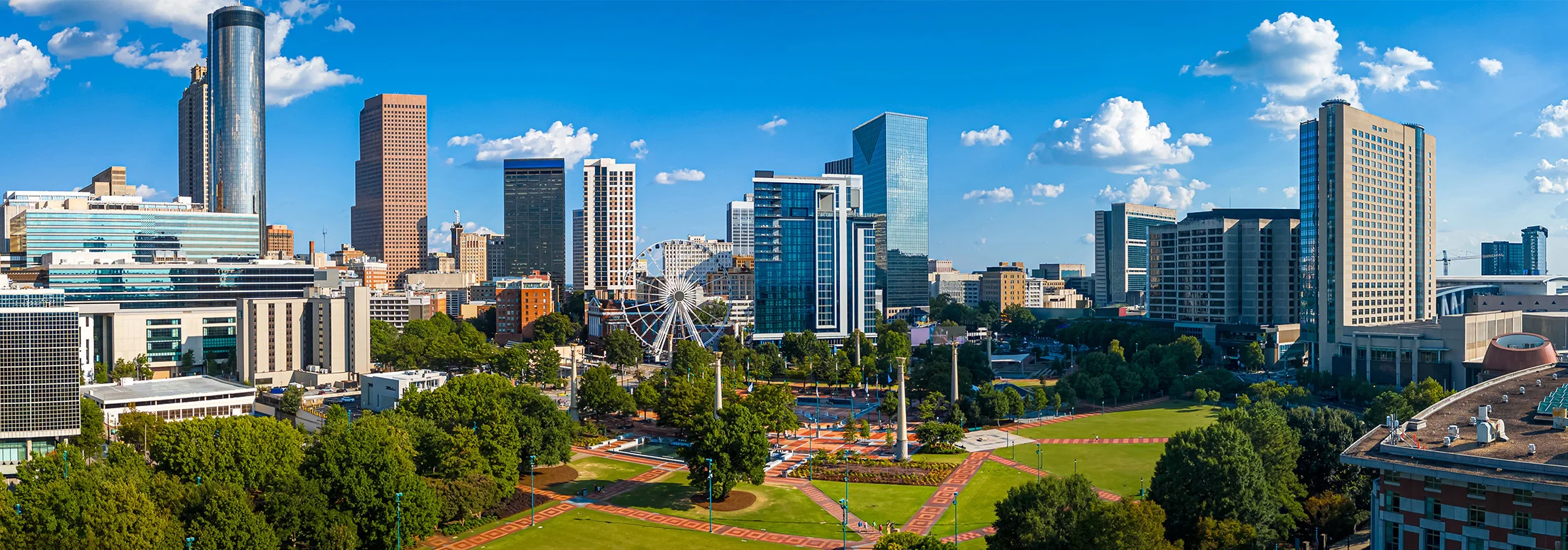 Atlanta skyline overlooking Centennial Olympic Park, featuring modern skyscrapers, green lawns, walking paths, and the Ferris wheel on a sunny day.