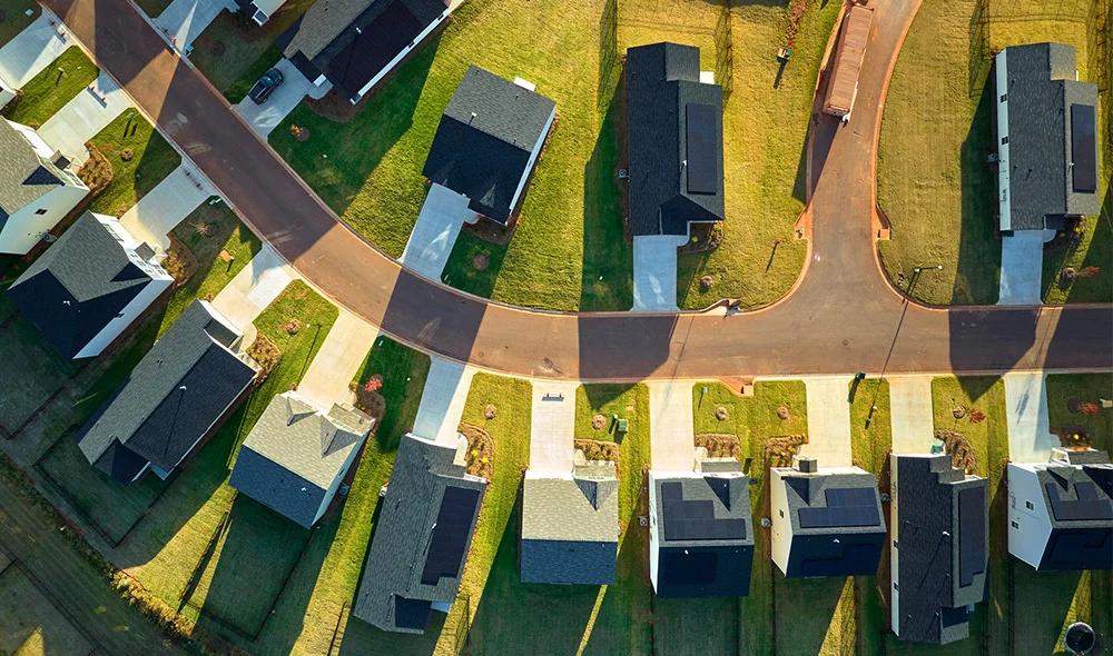 Aerial view of a suburban neighborhood showing rows of closely spaced houses with pitched roofs. The rooftops vary in color, including shades of gray, brown, green, and blue, creating a patchwork pattern. Each house has a small yard and driveways.