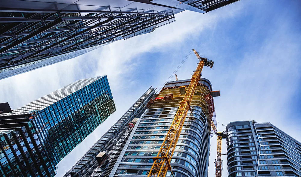 Upward-looking view of several modern high-rise buildings clustered closely together, creating a dramatic cityscape perspective. The buildings feature a mix of glass and steel exteriors with reflective windows and clean, vertical lines.