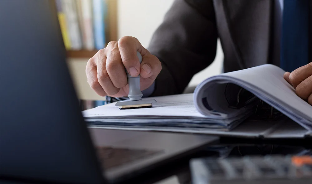 Close-up of a professional workspace showing a person in business attire writing with a pen on a document placed on a desk. A laptop is positioned to the left, partially open, with its screen facing away from the viewer.