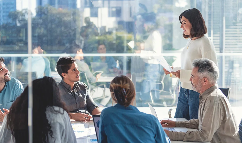 A group of professionals gathered in a modern office meeting room with large windows in the background. One individual stands at the front near a wall-mounted display screen, appearing to present or explain information facing the group.