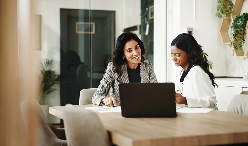 Two professionals collaborate at a desk in a bright office setting. One individual sits behind a laptop placed on a light-colored table, while the other stands beside the table and leans slightly inward, looking toward the screen. The workspace is clean.