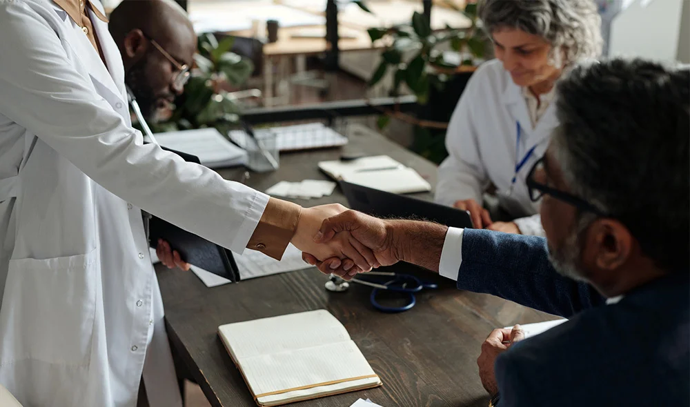 Close-up of a professional interaction in an office setting, showing two individuals in business attire reaching across a desk to shake hands. One individual extends a hand from the left side of the frame.