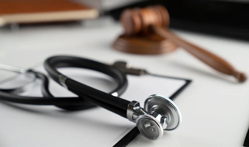Close-up of a stethoscope placed on a desk beside a white document. The stethoscope’s black tubing loops across the surface, with the metal chest piece resting near the center. In the softly blurred background, a wooden judge’s gavel is visible.