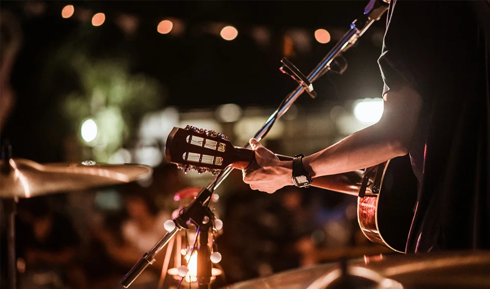 A small group of musicians performing on an outdoor stage at night, illuminated by warm hanging string lights overhead. Several individuals hold instruments while standing close together, with microphones and speakers visible around them.