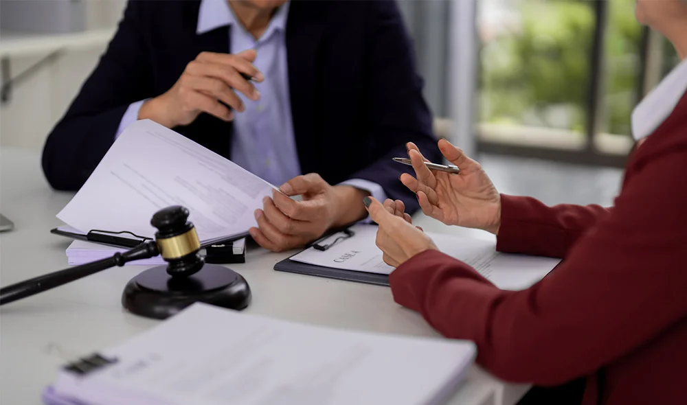 Close-up of a professional meeting at a desk, focusing on hands and paperwork. On the left side of the table, a person in a dark suit holds a pen above a document laid flat on the desk. Across from them, another person wearing a red top is visible.