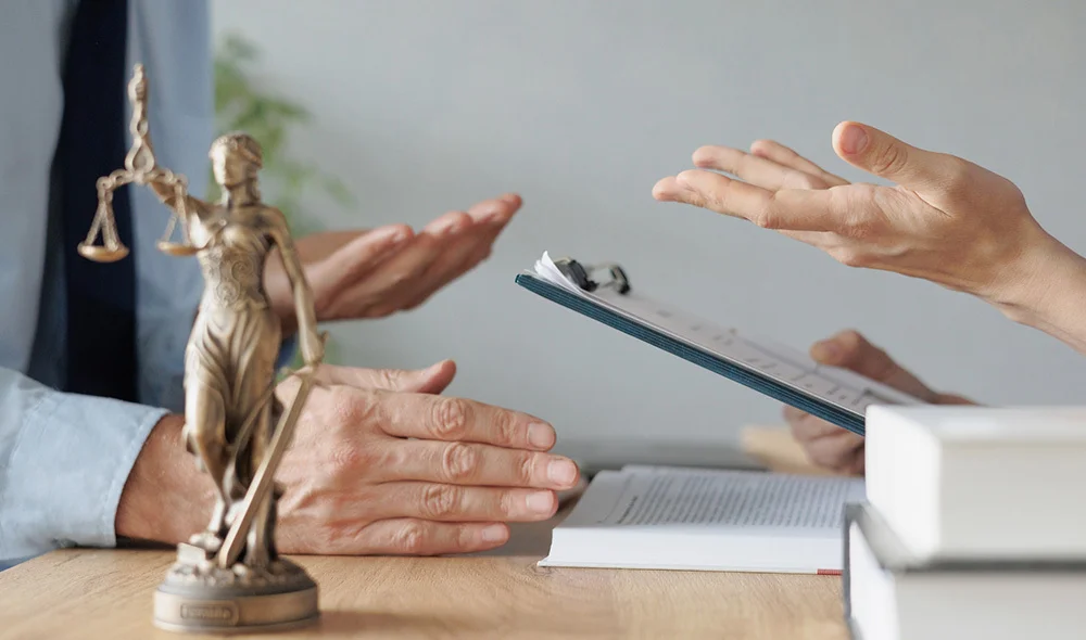 Close-up of a professional interaction at a desk, showing two individuals extending their hands toward each other in a handshake above a stack of documents. The handshake is centered in the frame, with both hands meeting over papers.