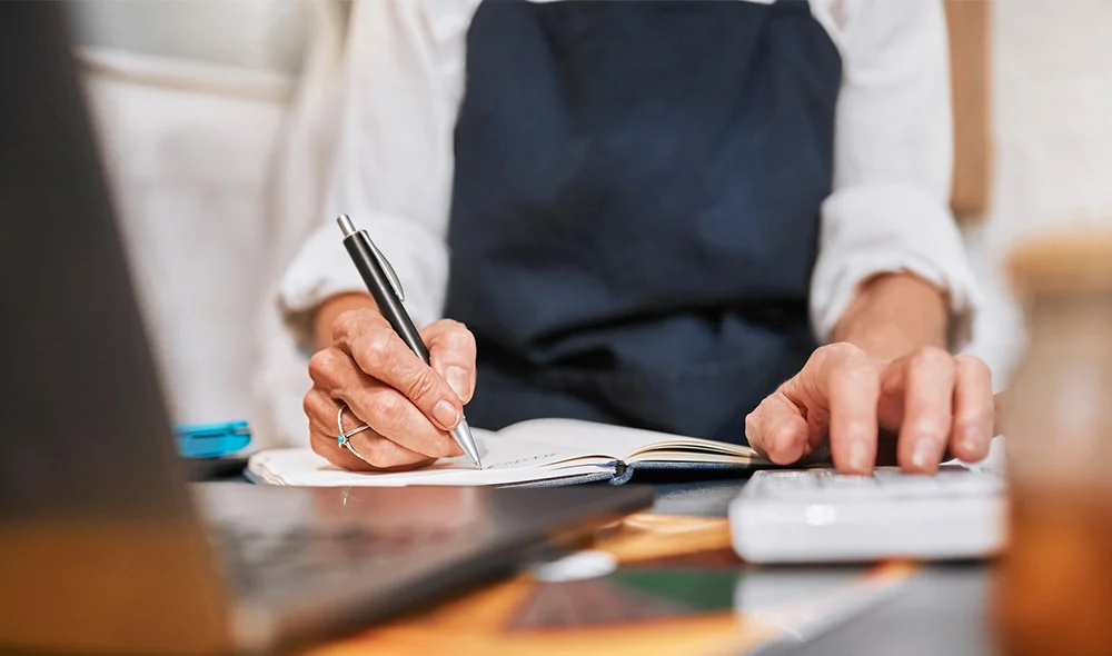 Close-up of a desk where a professional in business attire reviews documents. One hand holds a pen poised above a stack of papers, while the other hand steadies the documents. A wooden judge’s gavel and base sit prominently on the desk nearby.