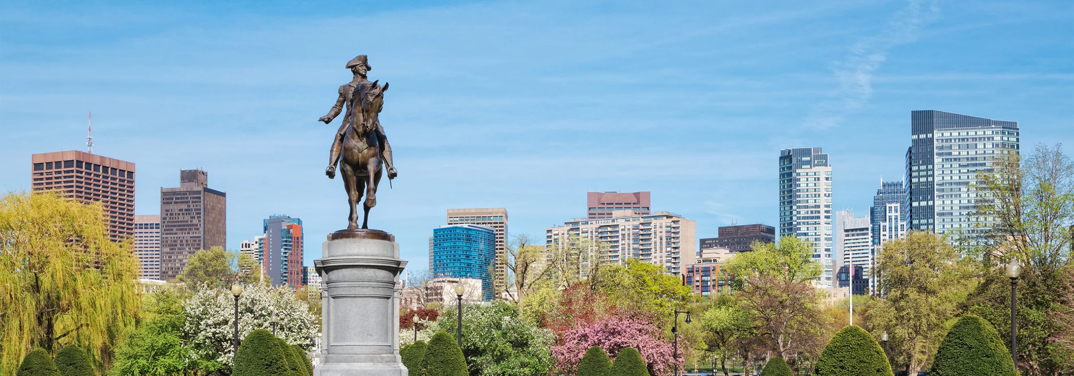 Paul Revere statue in a Boston Public Garden framed by blooming trees, with mid- and high-rise city buildings under a clear blue sky.