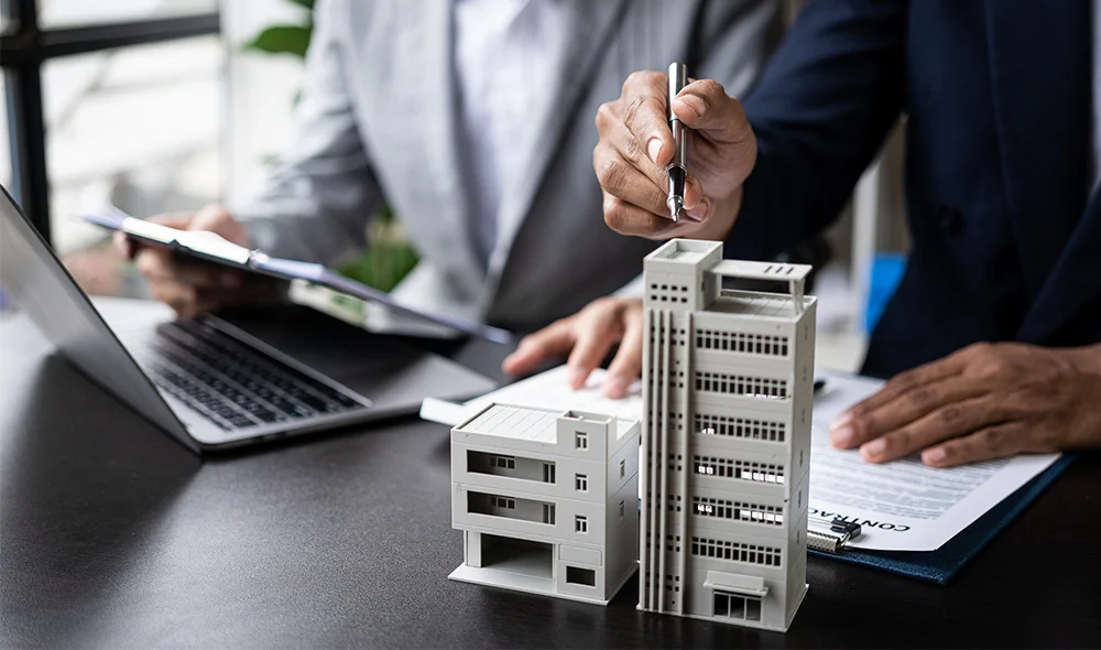 A close-up of a desk scene showing a person in business attire holding a pen and reviewing documents next to a laptop. Small architectural models of buildings are arranged on the table, suggesting a real estate or construction planning context.