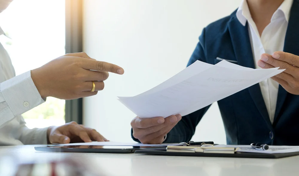 A close-up view of a business meeting at a desk where one person hands a document to another person. The receiving person holds the paper while a pen and additional documents rest on the table. Both individuals wear formal business attire.