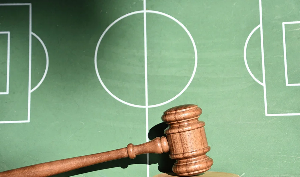An overhead view of a green soccer field diagram showing white outline markings for both goals, the center circle, and the midfield line. In the lower portion of the image, a wooden judge’s gavel rests on the field illustration.