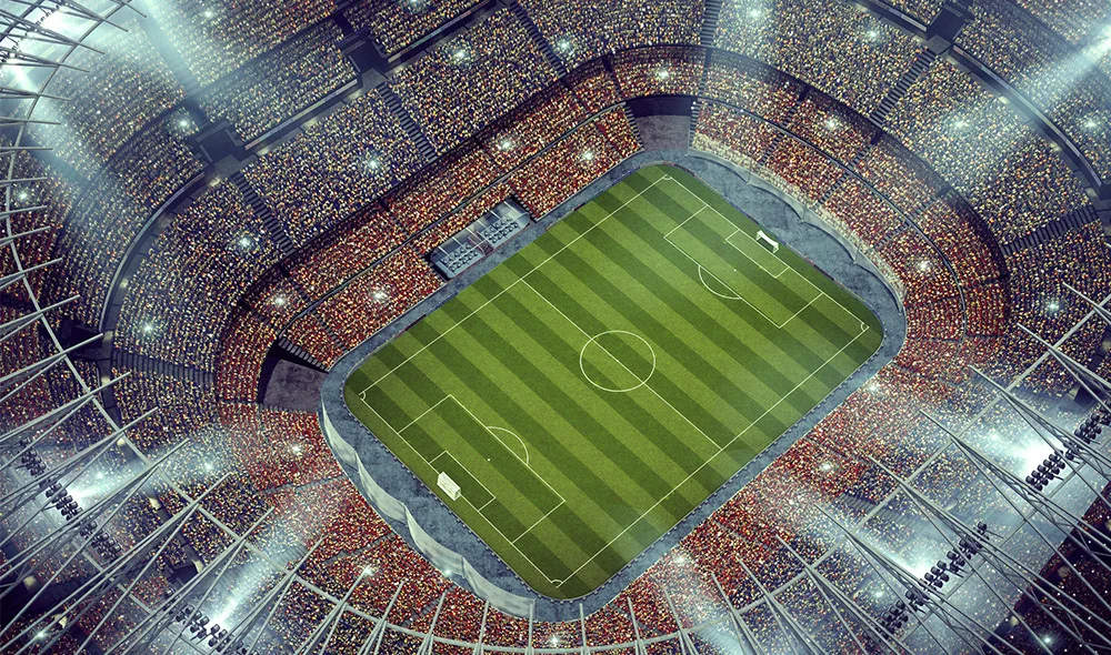 An aerial view of a large sports stadium with a bright green field at the center. The field is marked with clear lines, and rows of seating surround it on all sides. The stadium structure forms an oval shape, with streets, buildings, and parking.