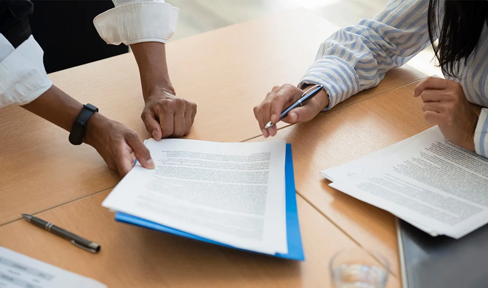 A top-down view of a collaborative workspace shows multiple people seated around a wooden table. Several hands reach toward documents and a tablet placed in the center, with laptops, notebooks, and papers spread across the table.