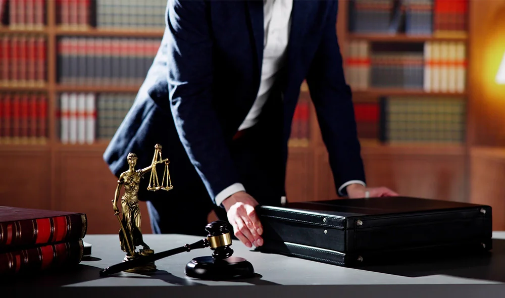 A business setting shows a person in formal attire pulling a small rolling suitcase while holding the handle of a briefcase. The individual is walking through an indoor area with warm lighting and a brick or tiled background.