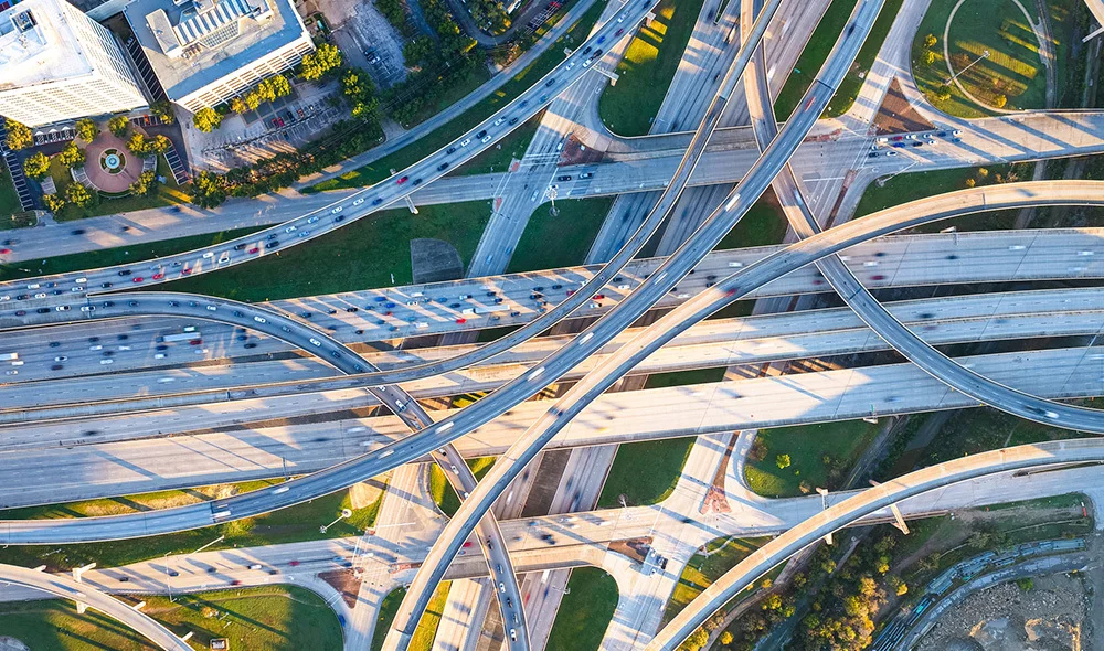 An aerial view of a complex highway interchange shows multiple elevated roads curving and intersecting in intricate patterns. Cars and vehicles are visible traveling along the lanes, with green patches of grass and landscaped areas filling the spaces.