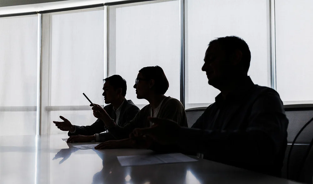 A silhouette scene of several people seated around a conference table in a modern office with floor-to-ceiling windows. One person gestures with a raised hand while others sit facing each other, suggesting an active discussion or meeting.
