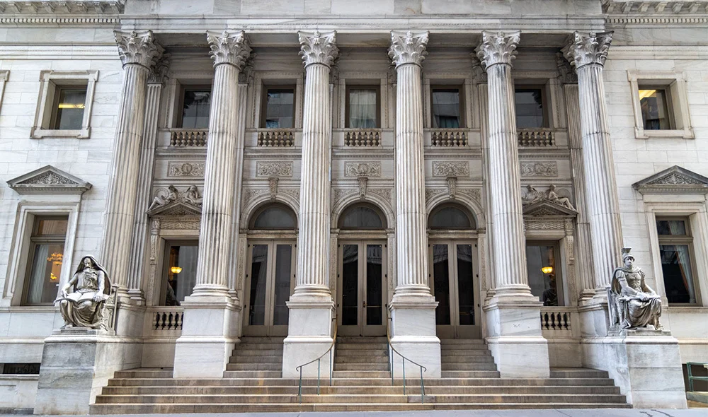 The front façade of a large neoclassical building with tall, evenly spaced stone columns supporting an elaborate entablature. Broad steps lead up to multiple grand entry doors, and the architecture features ornate detailing and a symmetrical design.