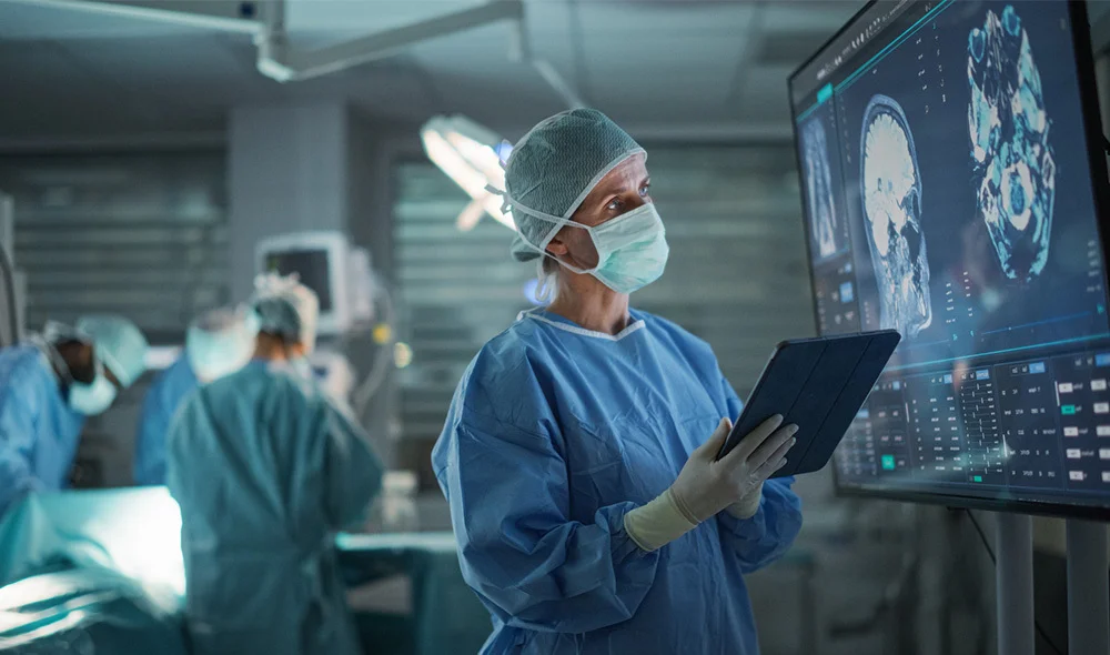 A high-tech workspace shows several individuals wearing lab coats, hair coverings, and protective gloves working in a clean, sterile environment. One individual in the foreground holds a digital tablet while looking toward a large transparent display.