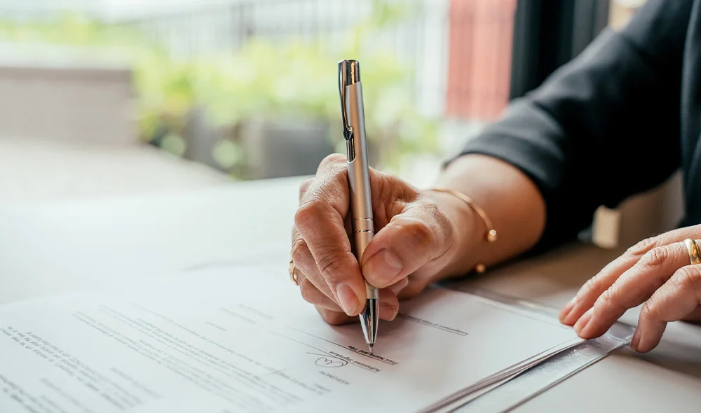 A close-up view of a person’s hand writing on a sheet of paper with a pen, resting on a smooth desk surface. The paper appears to contain printed text or forms, while additional documents and a folder are partially visible nearby.
