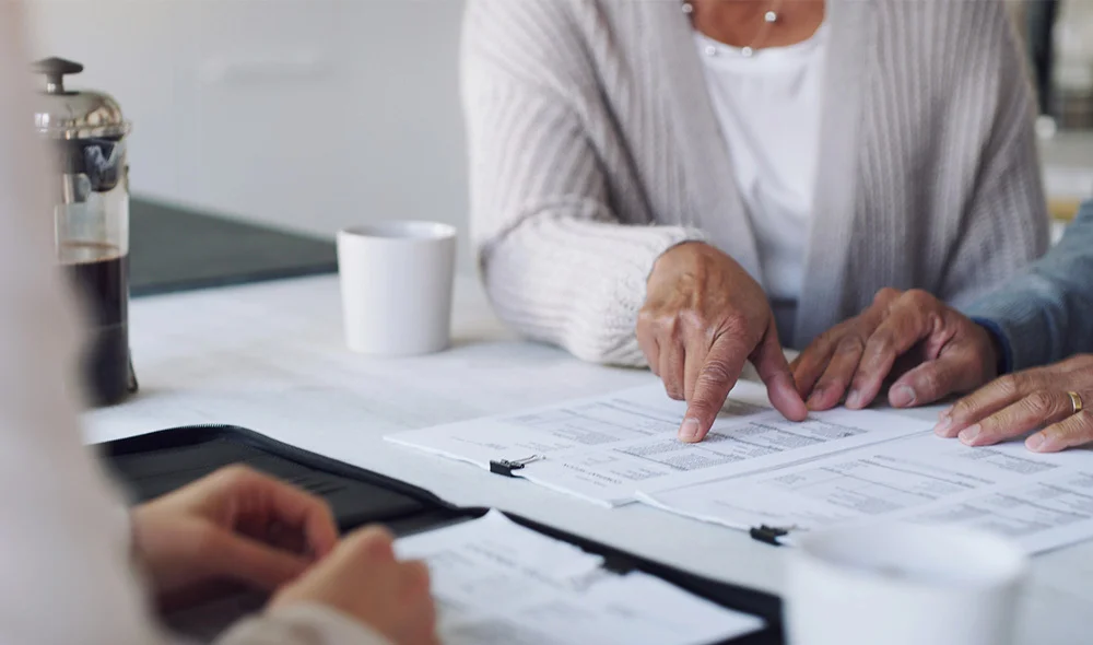 A close-up scene of a professional meeting at a desk shows two people reviewing paperwork together. One person wearing a light-colored sweater holds a pen and points to a document on a clipboard, while another person is across the table.