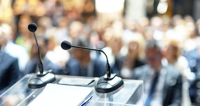 A shallow depth-of-field view of a conference or presentation setting shows a microphone on a stand and a pair of eyeglasses resting on a table in the foreground. In the blurred background, a large audience of people is seated, facing forward.