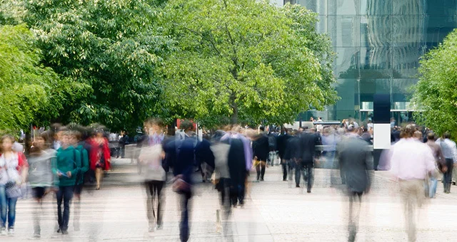 A busy outdoor urban plaza with numerous people walking in different directions, captured with motion blur. Trees with dense green foliage line the walkway, and modern glass buildings rise in the background, creating a contrast between greenery and city a
