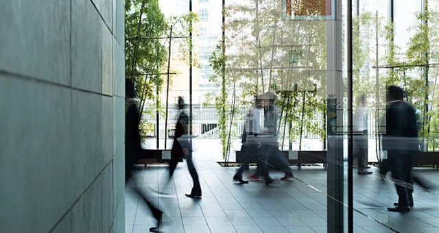 A modern office lobby with floor-to-ceiling glass walls and tall indoor plants arranged in rows. Several people in business attire walk through the space, captured with motion blur. Reflections from the glass surfaces mirror the movement, while natural li