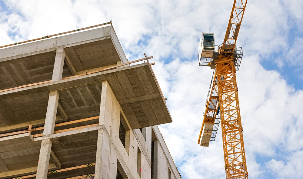 Wide-angle view of a construction site featuring a partially built concrete structure with multiple open floors and support columns. A tall yellow tower crane stands beside the building, extending into a partly cloudy sky, highlighting active development
