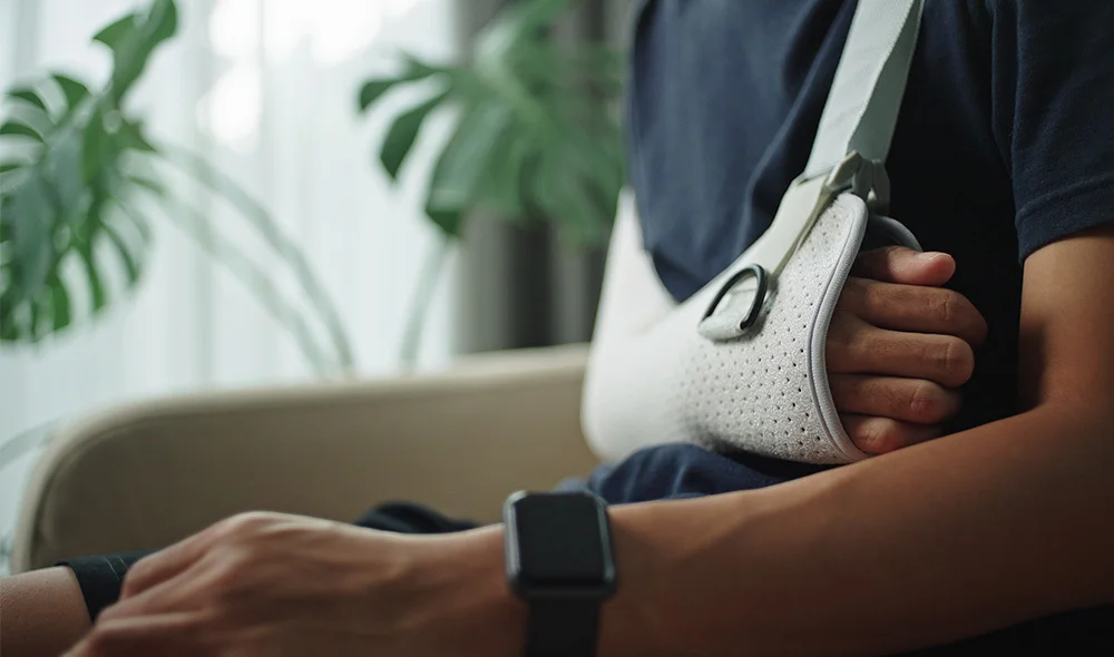 Close-up of a seated person wearing a white arm sling, with one hand resting inside the sling and the other on their lap. The person wears a smartwatch, and a leafy plant and soft interior lighting appear in the background, suggesting a comfortable indoor