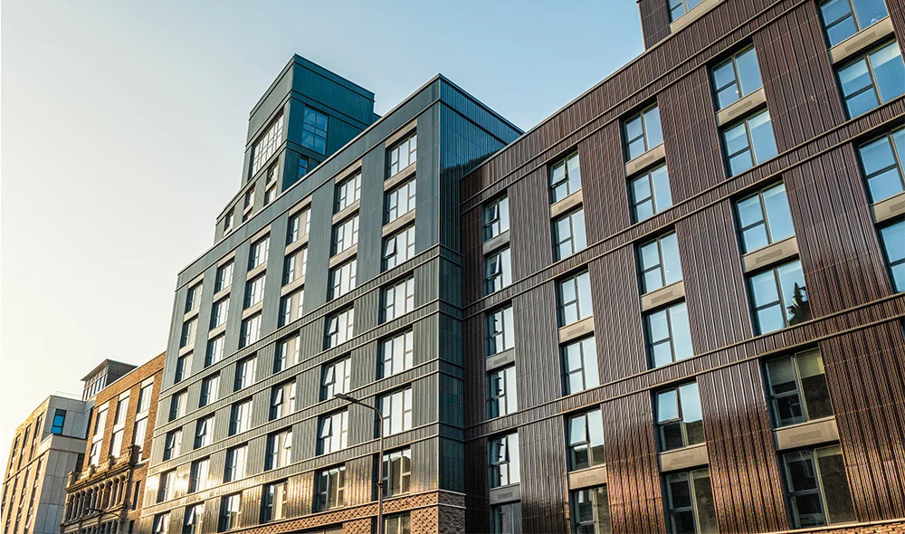 Modern mid-rise apartment or condominium building with a mix of brick and glass exterior, featuring large windows and varied facade textures. The structure rises several stories high under a clear sky, with neighboring buildings partially visible along th