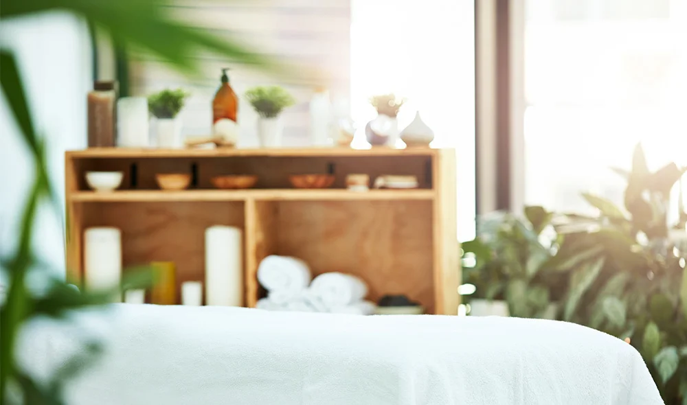 Bright, spa-like room with a neatly made massage table in the foreground. In the background, a wooden shelf holds rolled white towels, small bowls, and decorative items, while lush green plants and soft natural light from large windows create a calm, rela