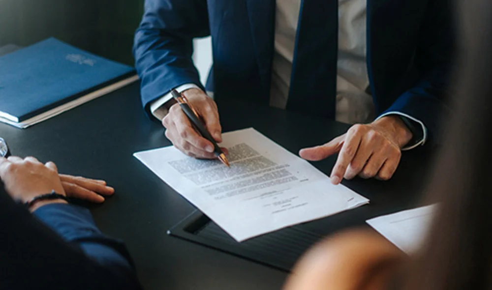 Close-up of a business meeting at a desk, where a person in a suit points to and marks a printed document with a pen while others review the paperwork nearby, suggesting a contract discussion or legal consultation.