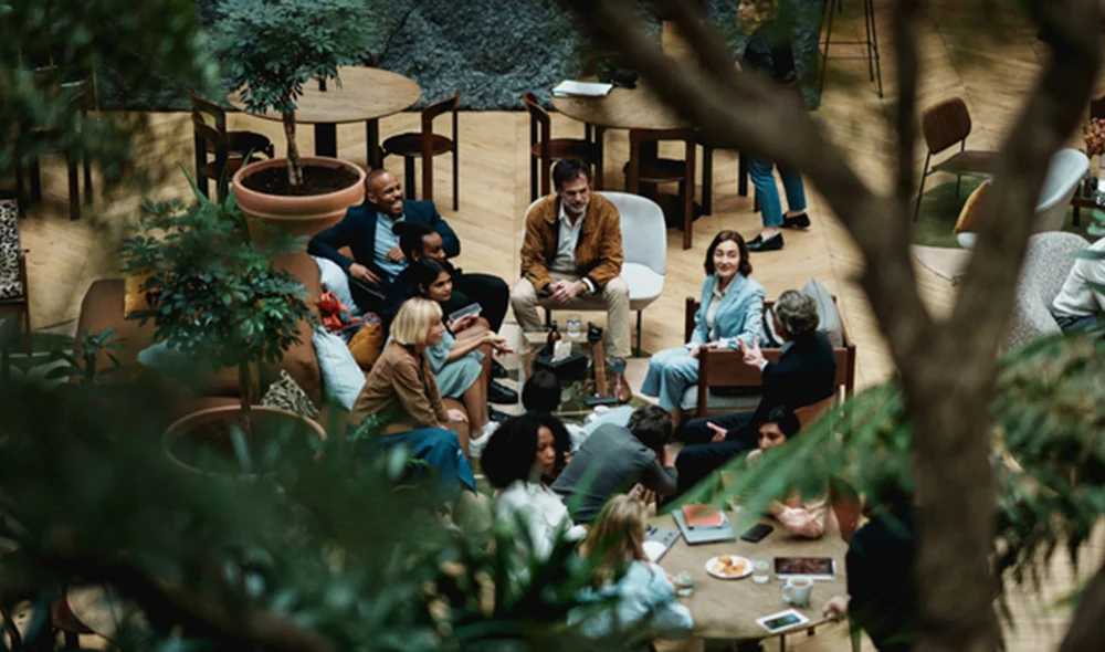Group of people gathered in a modern indoor courtyard, seated around tables and chairs in a casual meeting or discussion, surrounded by greenery and potted plants, viewed through tree branches in the foreground.